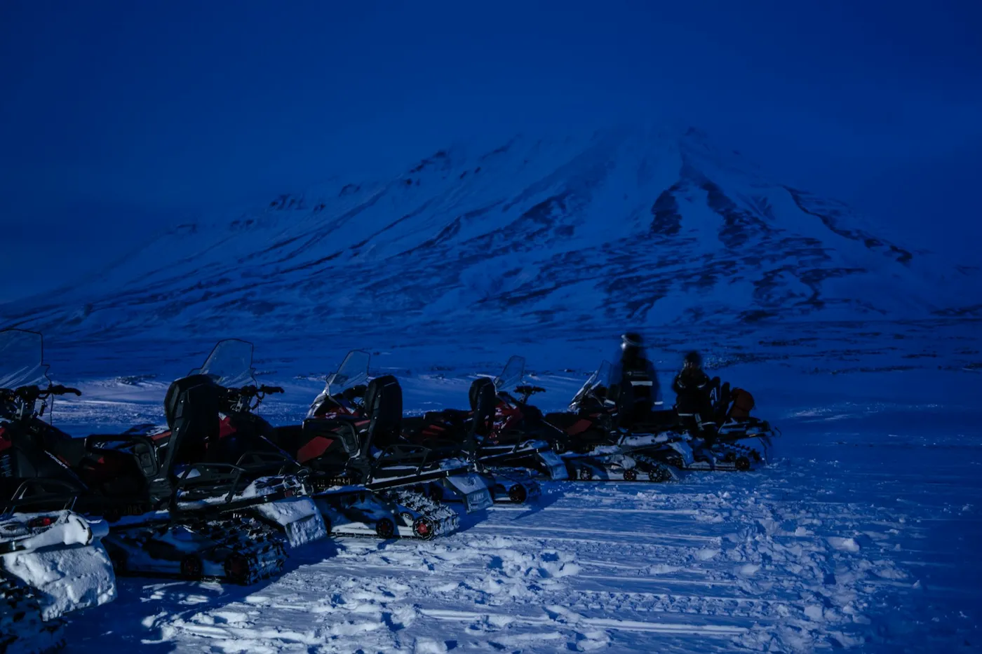 Una fila di motoslitte parcheggiate sulla neve battuta sotto una montagna innevata, tutta la scena immersa in una luce blu intensa