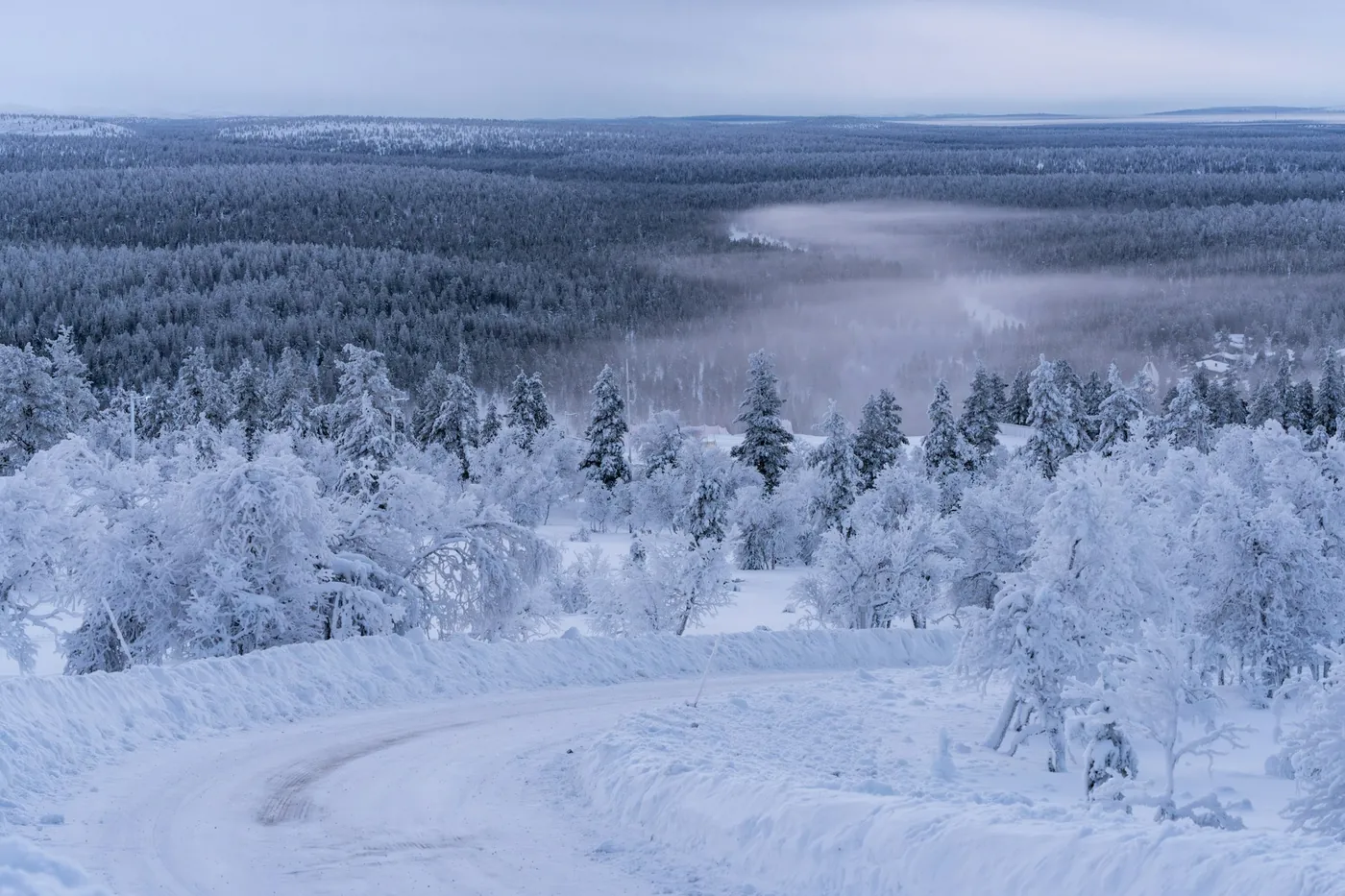 Une route enneigée descend en courbe entre des bouleaux chargés de givre, avec de la brume sur les vallées boisées au loin
