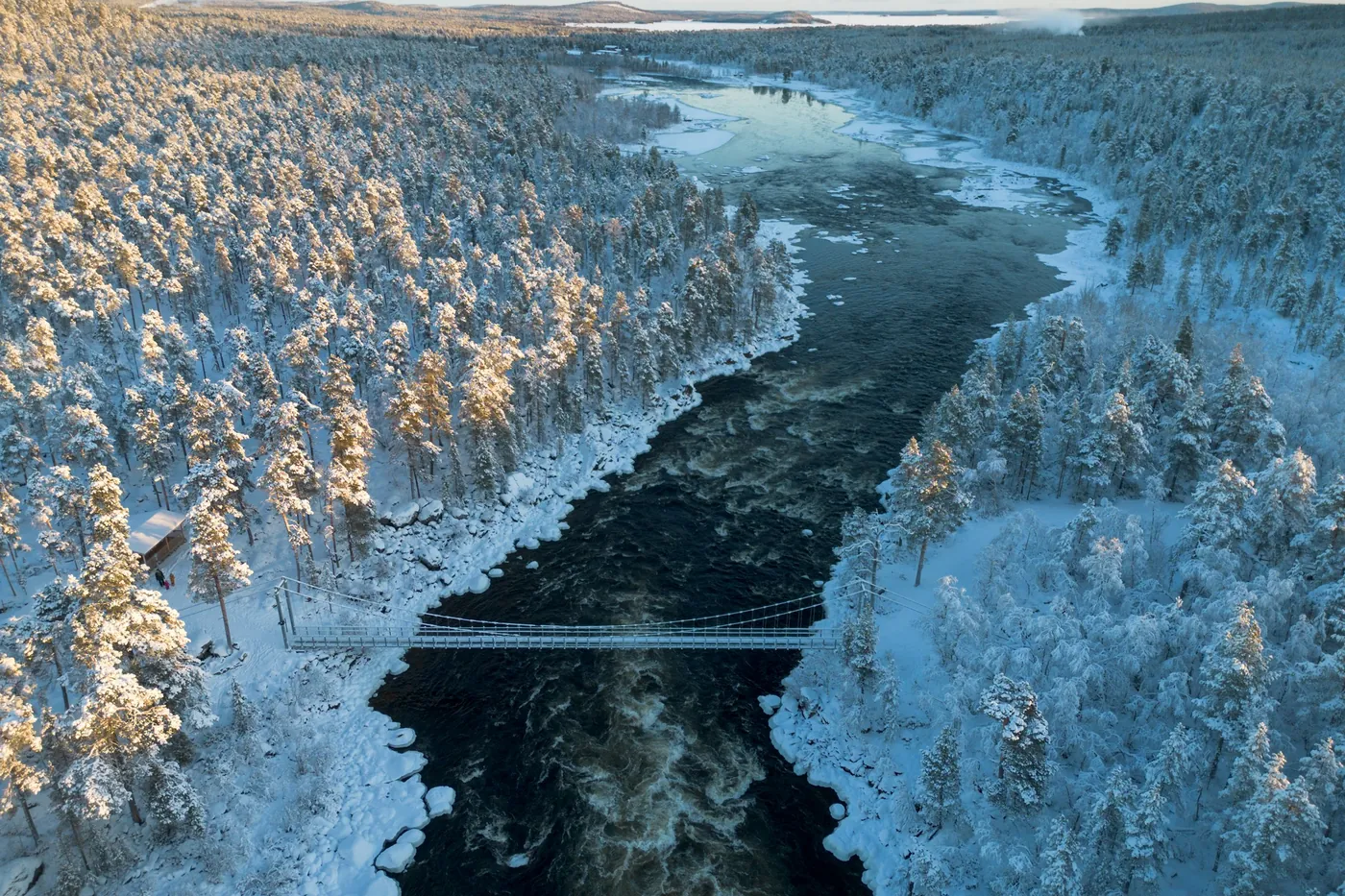 Aerial view of a suspension bridge crossing dark rapids between snow-covered pine forest lit by low sun, a frozen lake in the distance