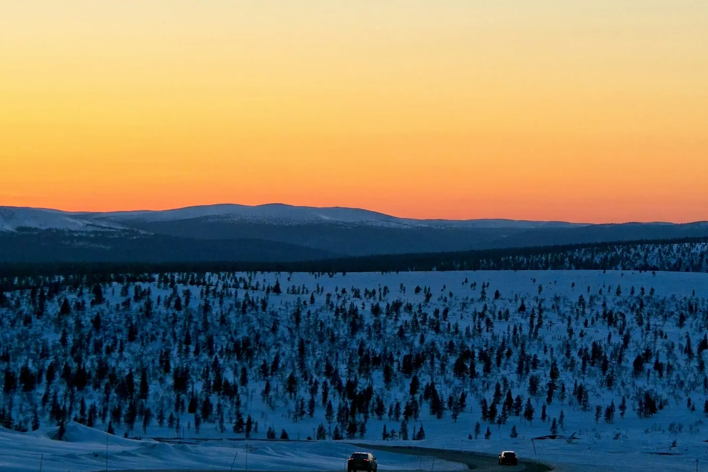 Cielo naranja sobre fells oscuros y nevados, con un bosque de pequeños abetos en la nieve y dos coches en una carretera en primer plano