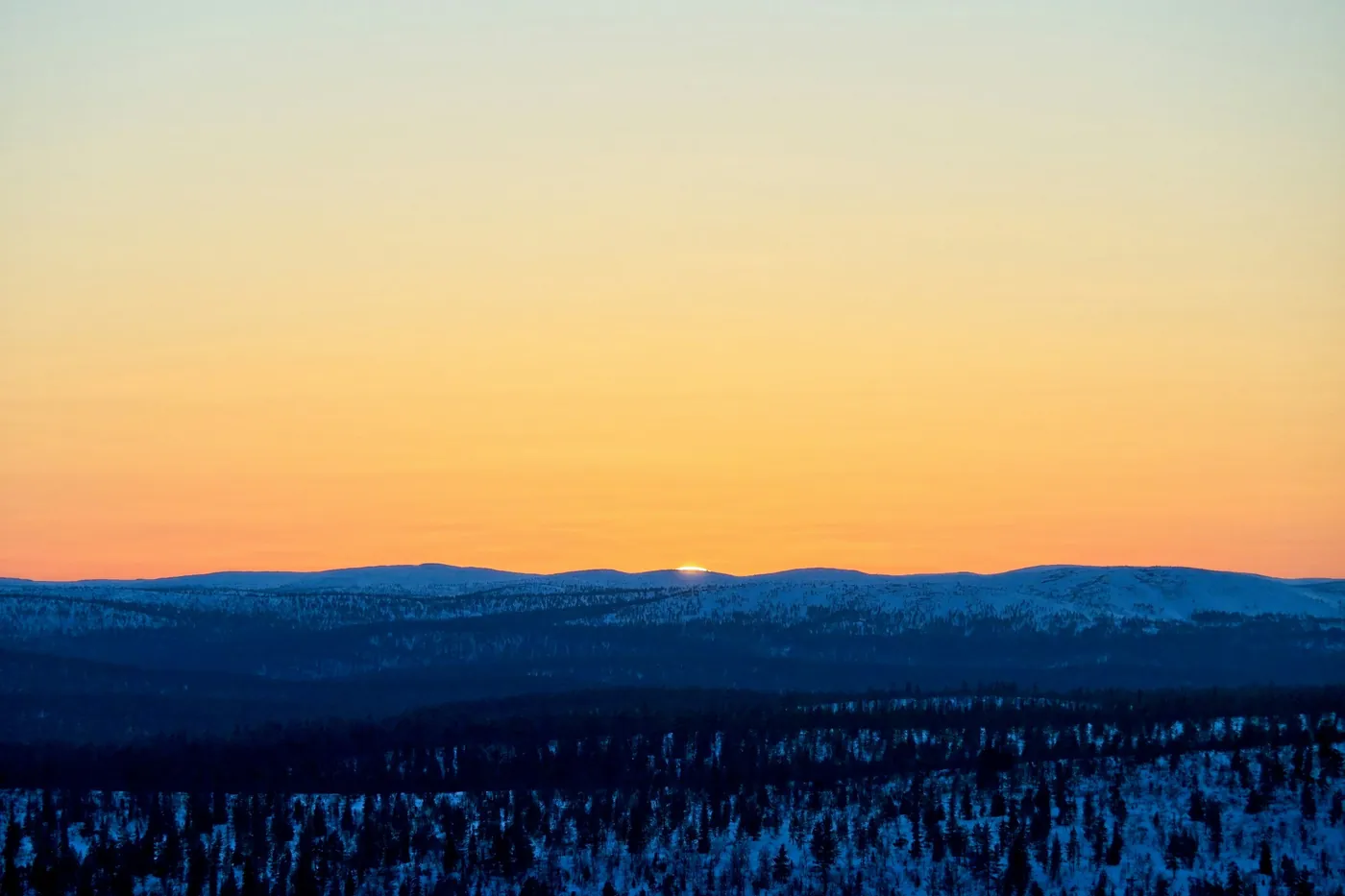Fells redondeados y nevados en el horizonte bajo un cielo naranja, con el sol rozando la cresta sobre un bosque oscuro