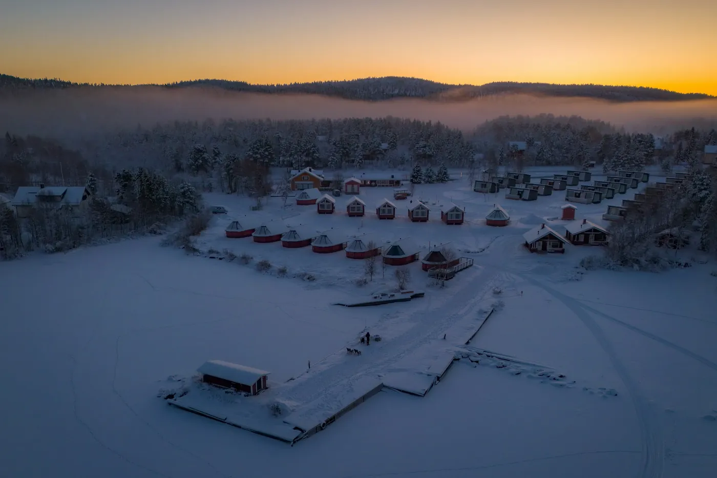 Luftaufnahme bei Sonnenaufgang: kleine runde Hütten und Häuschen an einem verschneiten Seeufer, ein Steg führt auf den zugefrorenen See, dahinter bewaldete Hügel im Nebel