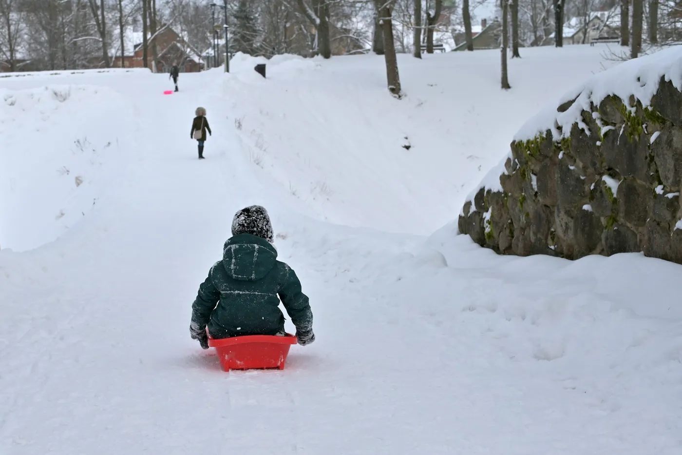 Ein kleines Kind in grüner Winterjacke und Strickmütze rutscht auf einem roten Plastikschlitten einen verschneiten Weg hinunter, weiter oben gehen zwei Personen