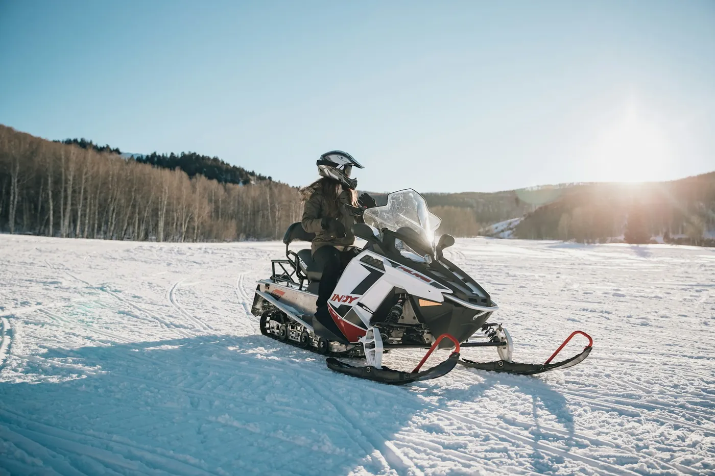 Ein Fahrer mit Helm sitzt auf einem weiß-roten Schneemobil auf einem verschneiten Feld, tiefe Sonne hinter kahlen Bäumen